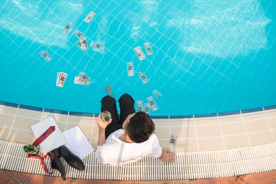 aerial of man sitting on pool edge with money floating in the pool